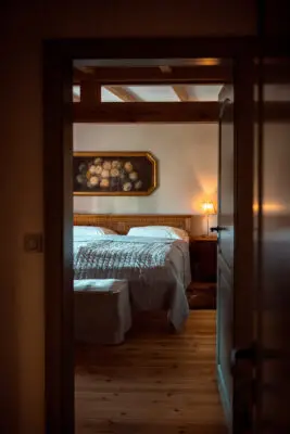 Blick durch eine offene Tür in ein gemütlich eingerichtetes Schlafzimmer mit Holzbalkendecke, Doppelbett und warmem Lampenlicht.